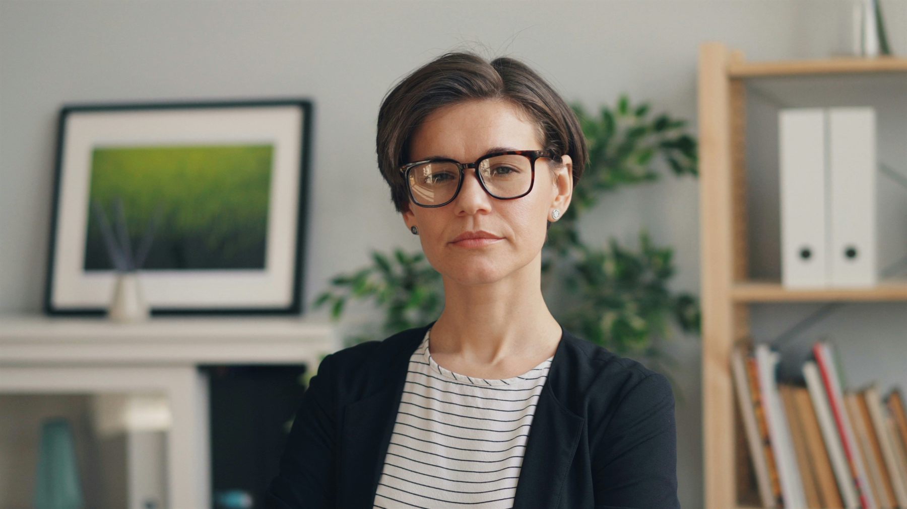A professional woman in a blazer and glasses looking at the camera in a bright office